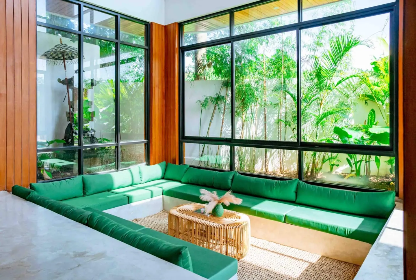 Sunken lounge conversation pit with emerald cushions, teak ceiling detail and full-height black-framed glass onto a bamboo garden